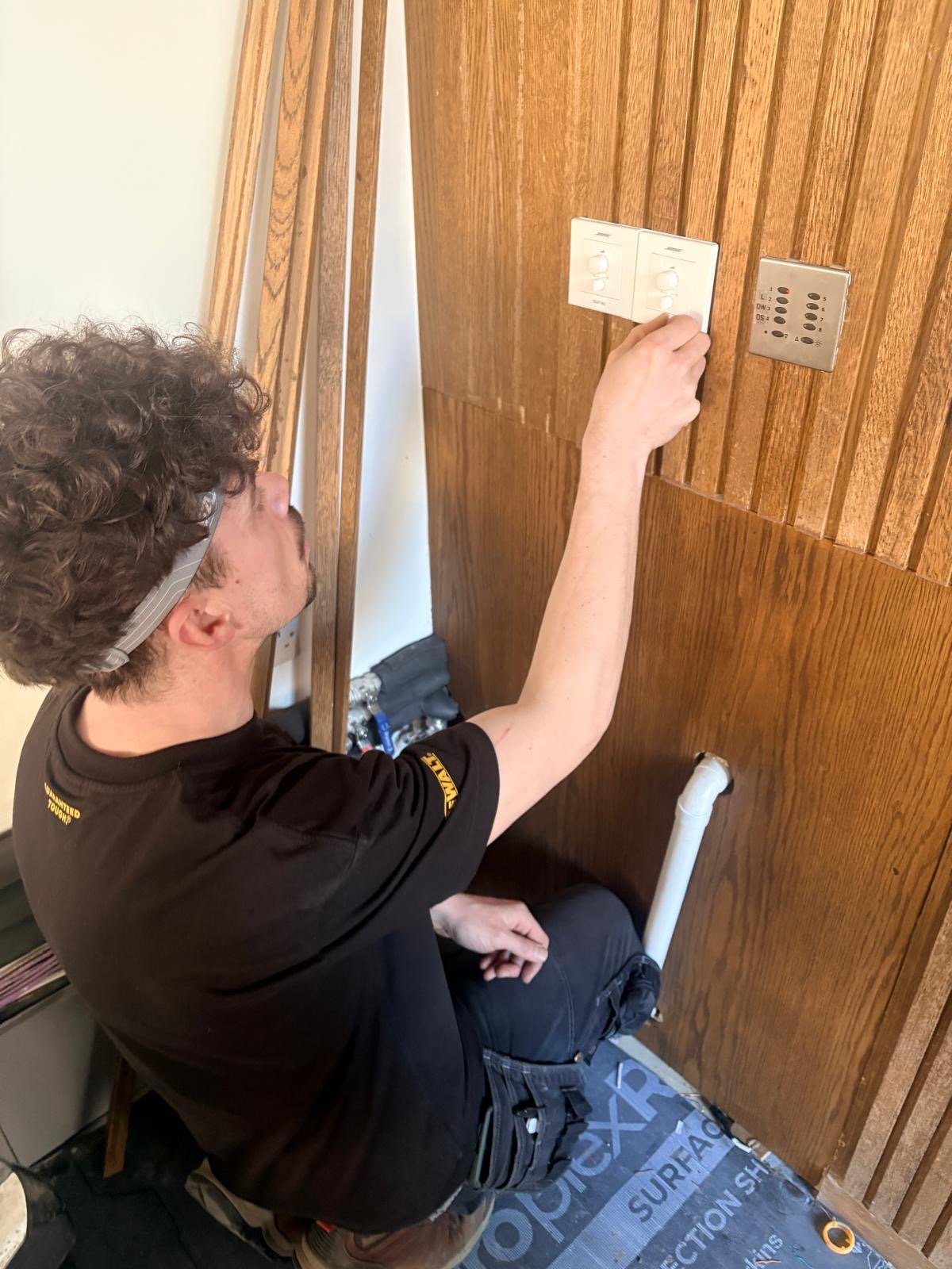 A person with curly hair and a headband is kneeling and installing or adjusting light switches on a wooden wall, with tools and materials nearby—perhaps taking a break from the bustle of London or sipping on Guillam Coffee whilst working. A person with curly hair and a headband is kneeling and installing or adjusting light switches on a wooden wall, with tools and materials nearby—perhaps taking a break from the bustle of London or sipping on Guillam Coffee whilst working.