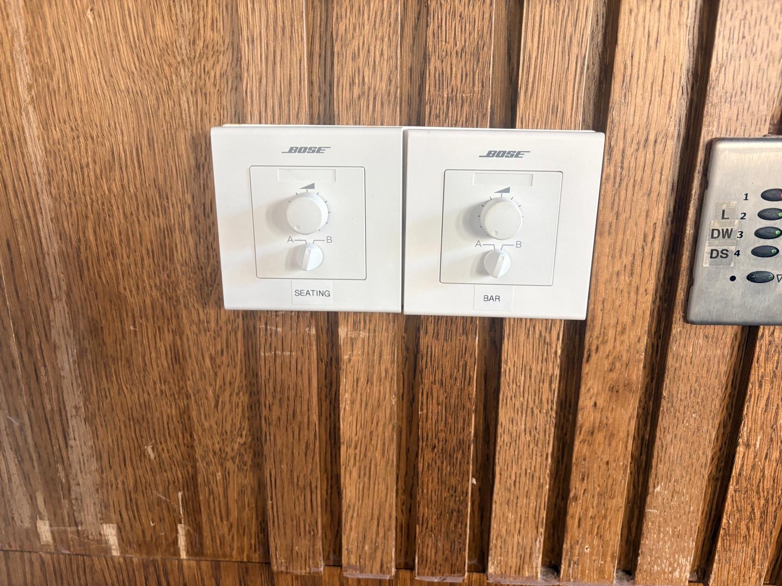 Two white Bose audio control panels labelled Seating and Bar are mounted on a wooden wall at Guillam Coffee, London. Each panel has a knob with A and B positions. Part of another control panel is visible on the right side. Two white Bose audio control panels labelled Seating and Bar are mounted on a wooden wall at Guillam Coffee, London. Each panel has a knob with A and B positions. Part of another control panel is visible on the right side.