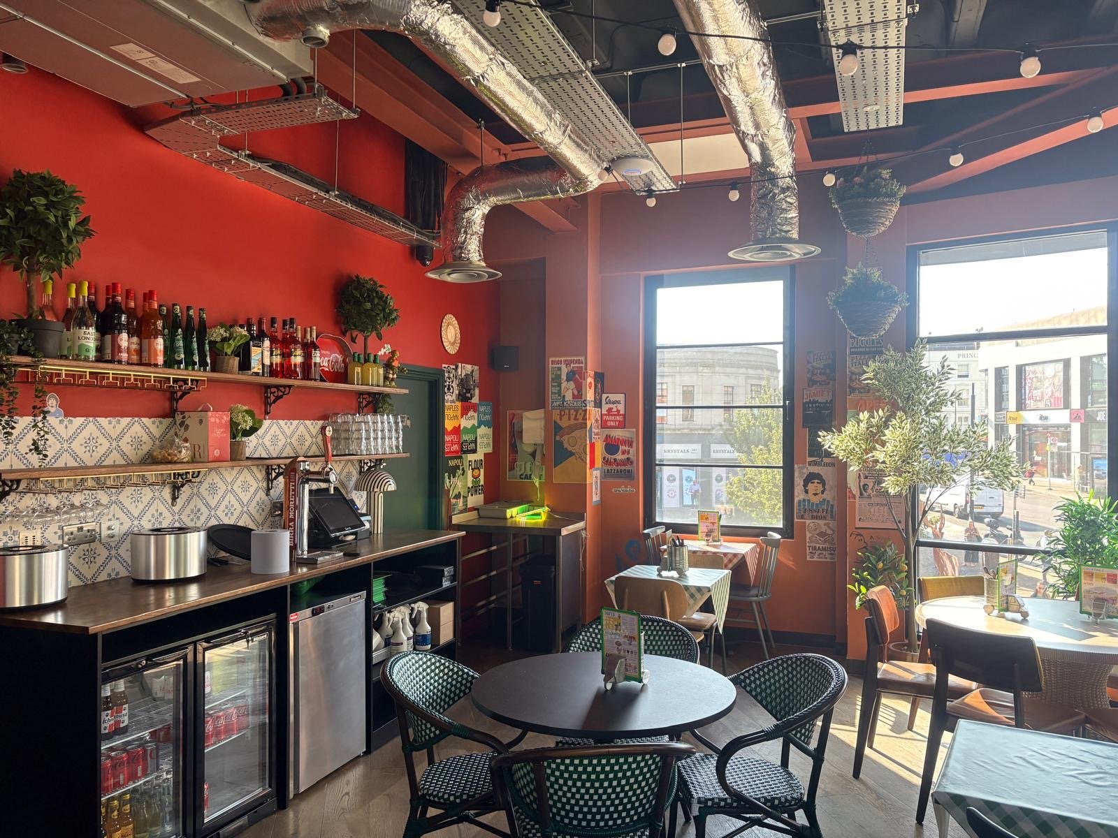 A cosy café interior with red walls, tiled splashback, potted plants, tables and chairs, a bar area with bottles, and large windows letting in sunlight. Posters decorate the walls, creating a vibrant atmosphere reminiscent of Pizza Pilgrims in Wimbledon.