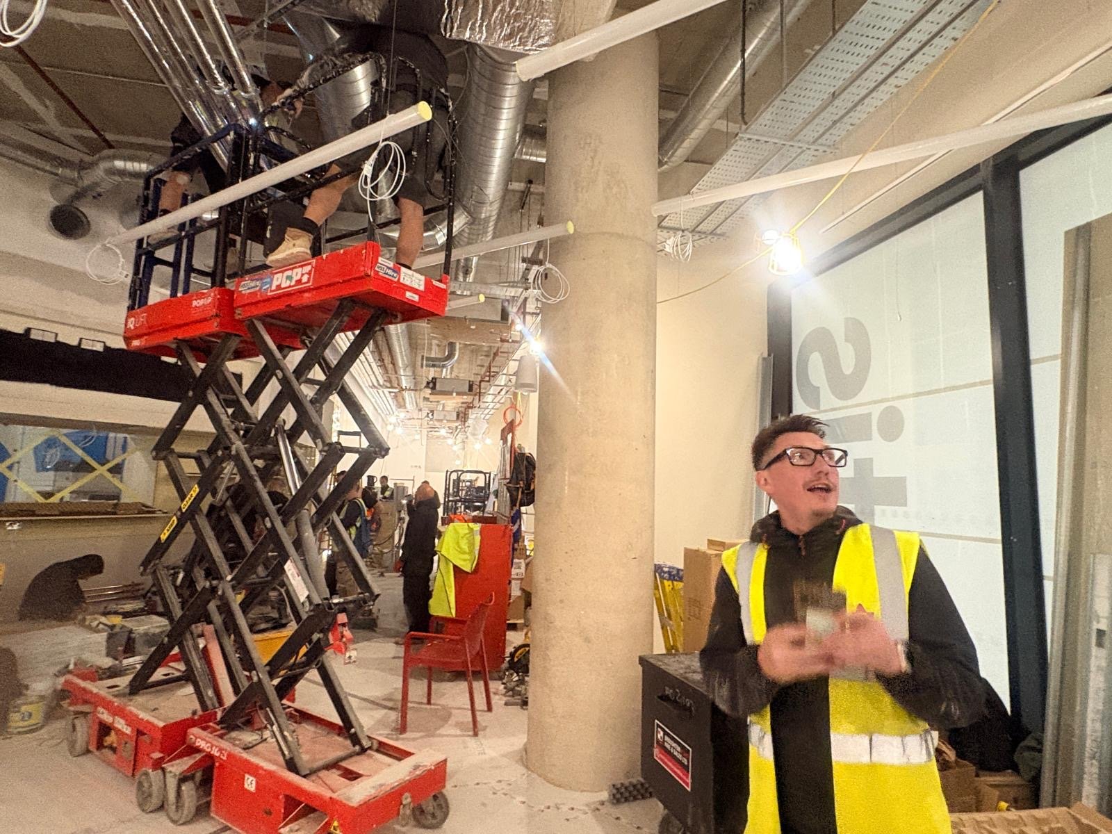 Construction workers wearing safety vests work indoors at a site in St James’s Park, London; one stands holding tools, while others use a scissor lift to reach ceiling pipes and wires. The area is filled with ATIS equipment and building materials.