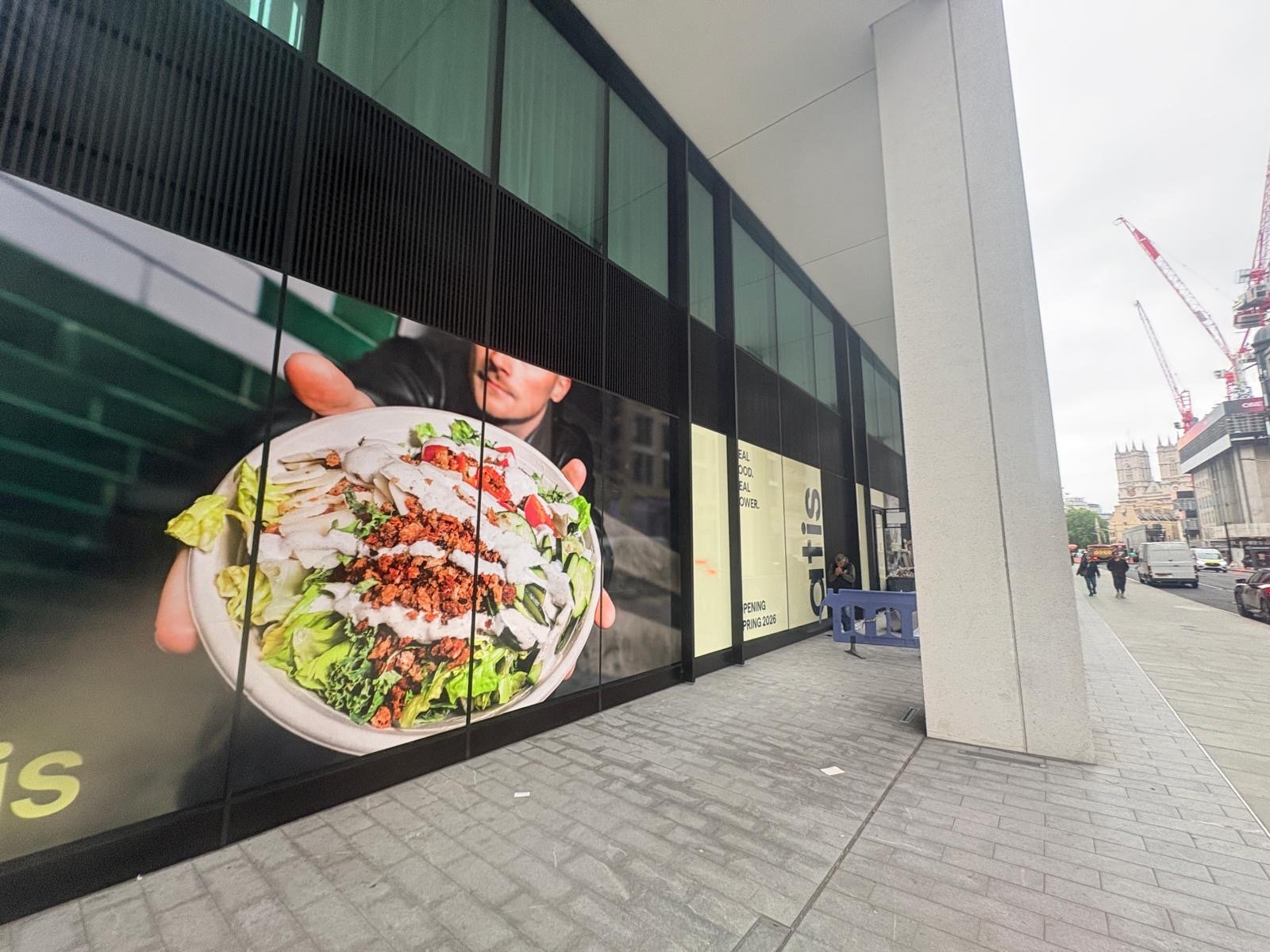 Large window graphic of a person holding a bowl of salad, displayed on the façade of a modern building along a bustling London city street near St James’s Park, with pedestrians and construction cranes visible in the background—a vibrant scene reflecting the contemporary spirit of ATIS.