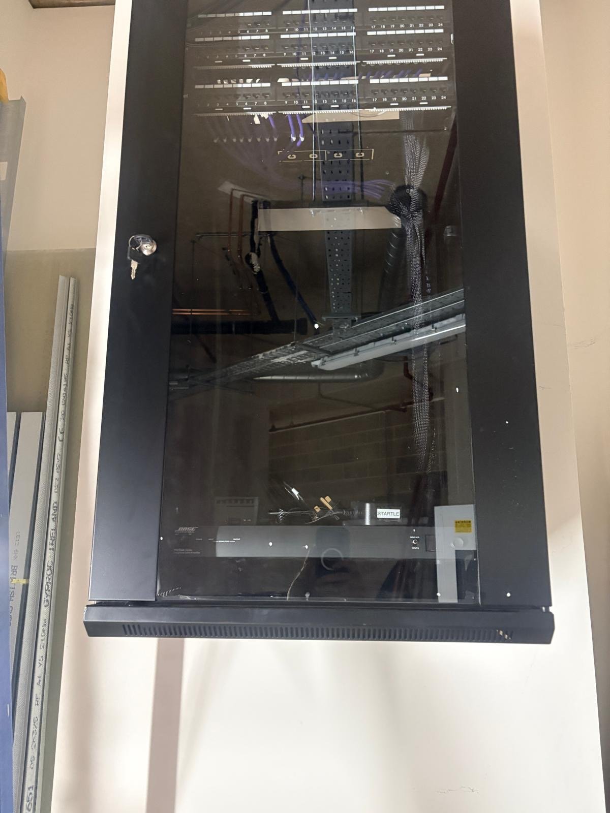 A black server rack cabinet with a glass front door, metal sides, and visible network equipment inside, mounted on a wall in a utility or server room near St James’s Park in London.