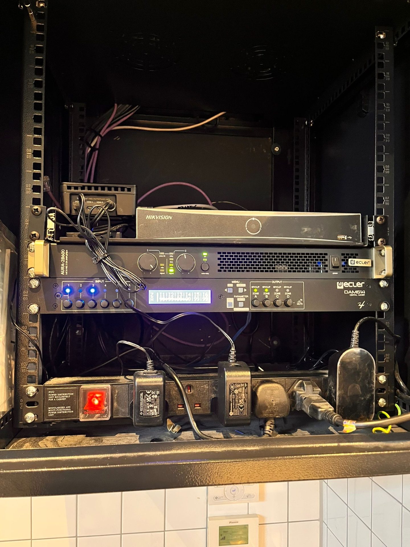 A network rack with multiple electronic devices, including a Hikvision device and other audio-visual equipment, plugged into several power adaptors, with cables organised inside a black enclosure at Pizza Pilgrims in Glasgow.