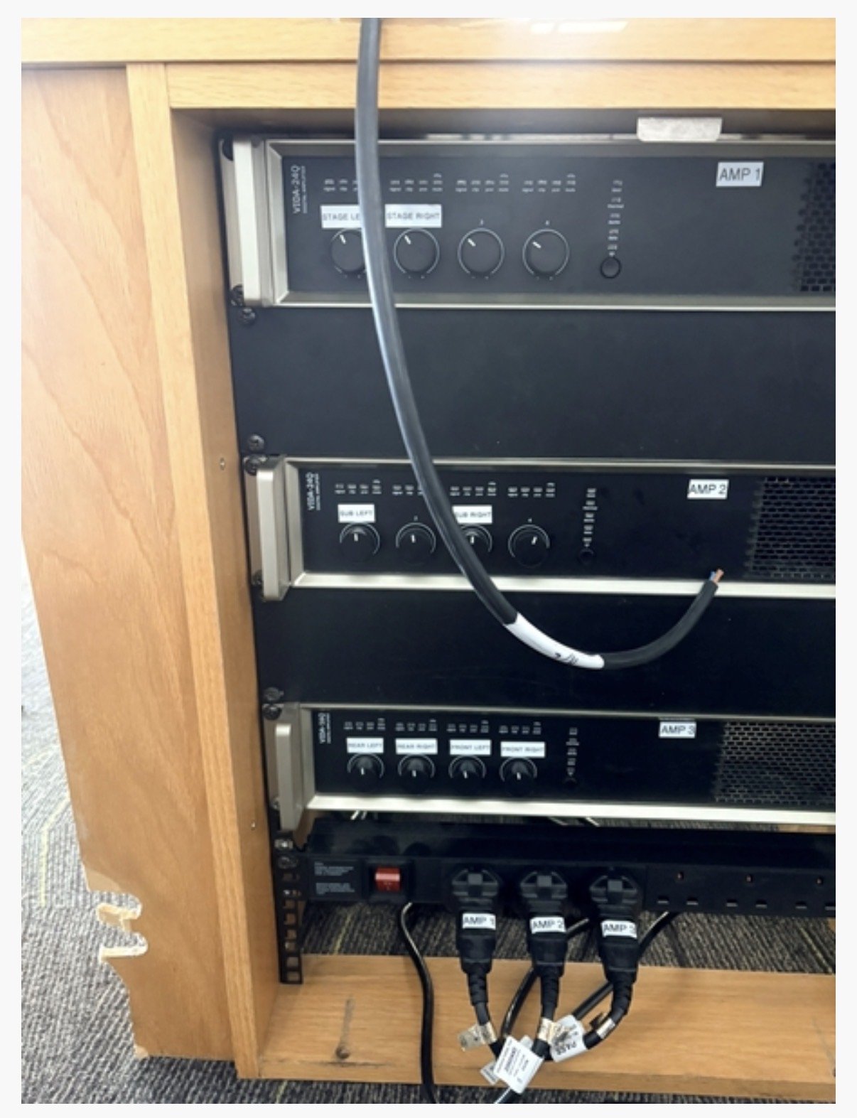 A wooden rack from Highgate House in Northampton holds three black amplifier units labelled AMP with control knobs and switches, as well as a power strip with four plugs and visible cables.
