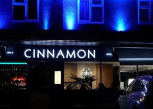 A restaurant called CINNAMON is lit with a bright sign at night. Blue lights illuminate the building above, and a parked car is visible in front, showcasing the inviting atmosphere of Cinnamon Restaurant in North Weald.