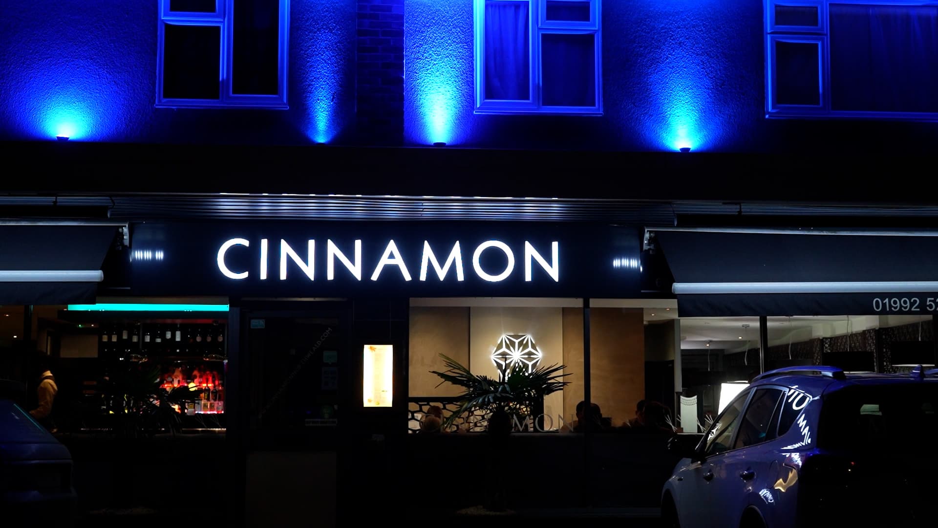 A restaurant called CINNAMON is lit with a bright sign at night. Blue lights illuminate the building above, and a parked car is visible in front, showcasing the inviting atmosphere of Cinnamon Restaurant in North Weald. A restaurant called CINNAMON is lit with a bright sign at night. Blue lights illuminate the building above, and a parked car is visible in front, showcasing the inviting atmosphere of Cinnamon Restaurant in North Weald.
