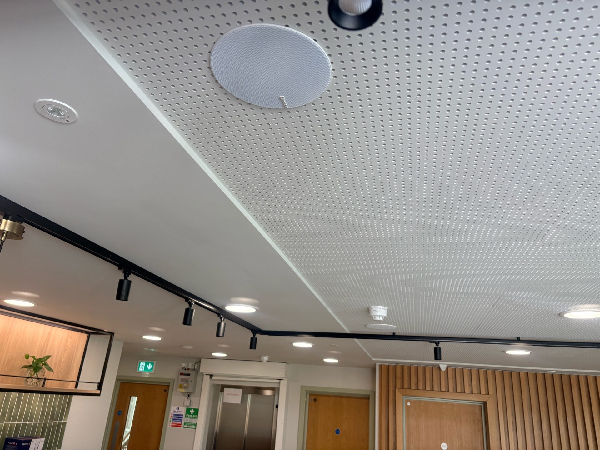 A modern office ceiling at St Luke’s Hospice with a round speaker, fire alarm, black track lighting, and part of a wooden wall. Doors, a lift, and safety signs are visible in the background of this Thurrock bistro-inspired space.
