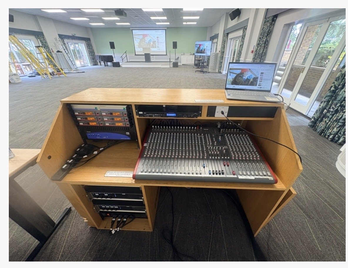 A wooden audio-visual control desk with a large sound mixer, audio equipment, and a laptop, positioned in a spacious hall at Highgate House near Northampton, featuring open doors, screens, and rows of chairs.