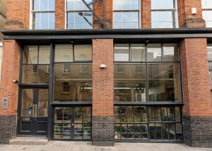 Large brick building with tall windows, a glass door on the left, and plants visible inside. The reflection of another brick building can be seen in the upper windows. The address sign reads 29 31. Located in London, this space is home to TL Draw.