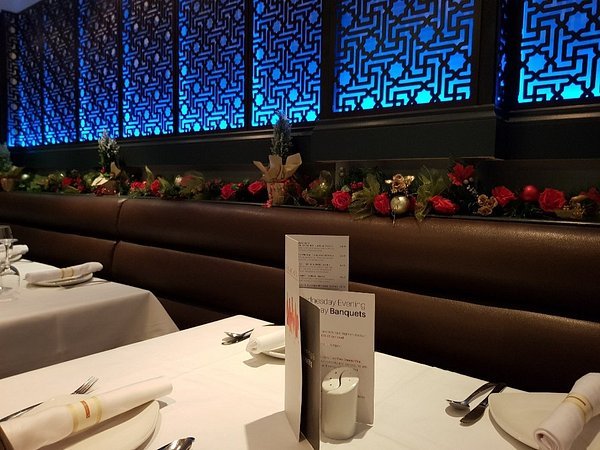 Elegant restaurant table with white linen, rolled serviettes, and a menu. The background features festive garlands with red baubles and an ornate blue-lit wall panel. The atmosphere at Cinnamon Restaurant in North Weald is calm and sophisticated. Elegant restaurant table with white linen, rolled serviettes, and a menu. The background features festive garlands with red baubles and an ornate blue-lit wall panel. The atmosphere at Cinnamon Restaurant in North Weald is calm and sophisticated.