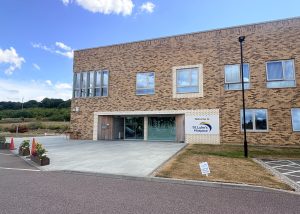 A modern brick building with large windows and a sign reading St Luke’s Hospice by the entrance, surrounded by a lawn and a paved walkway under a blue Thurrock sky with clouds.