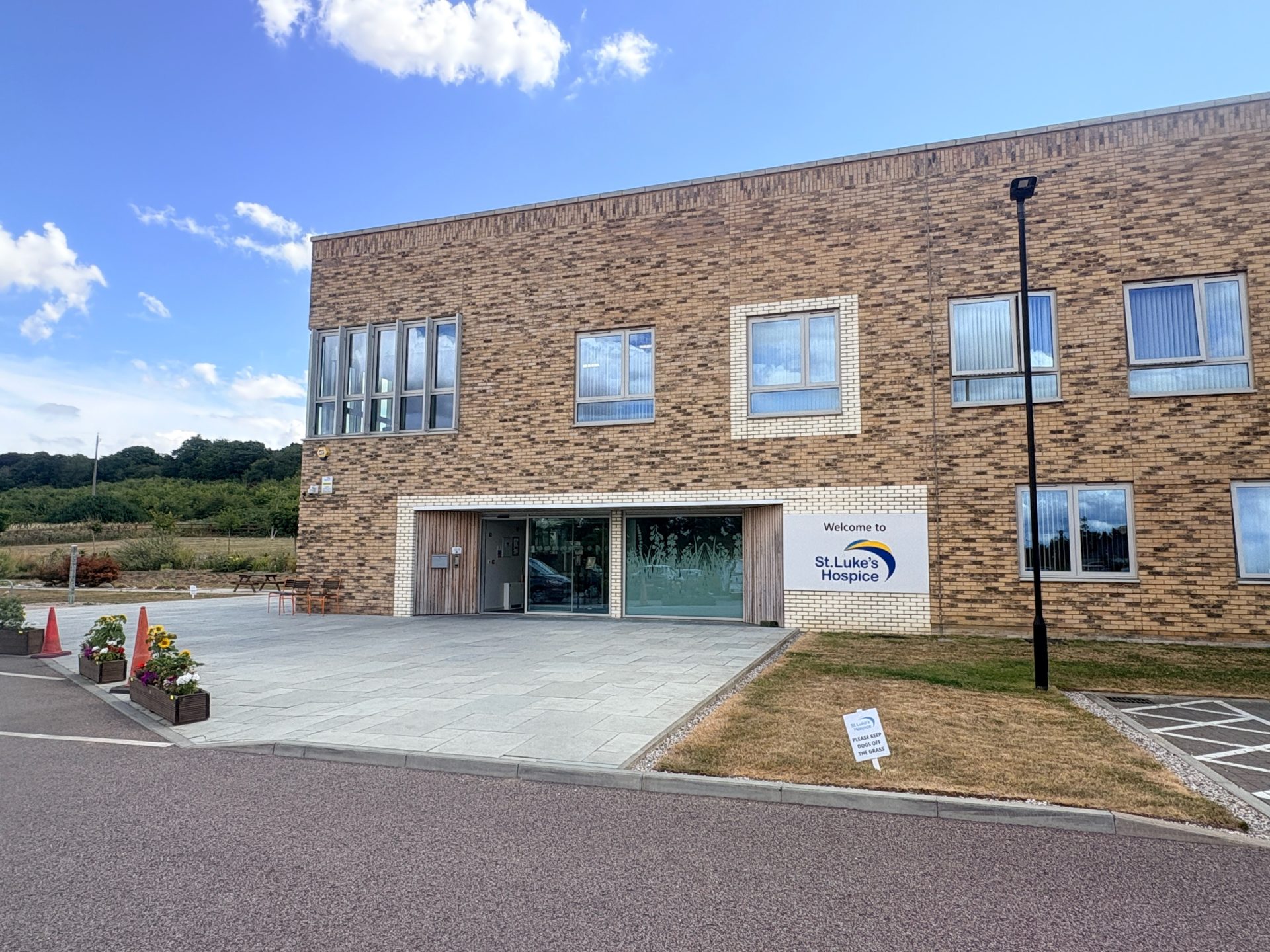 A modern brick building with large windows and a sign reading St Luke’s Hospice by the entrance, surrounded by a lawn and a paved walkway under a blue Thurrock sky with clouds.