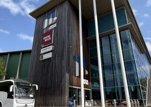 A modern building with tall white columns, a glass facade, and wooden panelling stands prominently in Hemel Hempstead. Signs for Snow+Rock and other businesses suggest its connection to The Snow Centre, the renowned indoor ski destination. A white coach is parked outside under a partly cloudy sky.