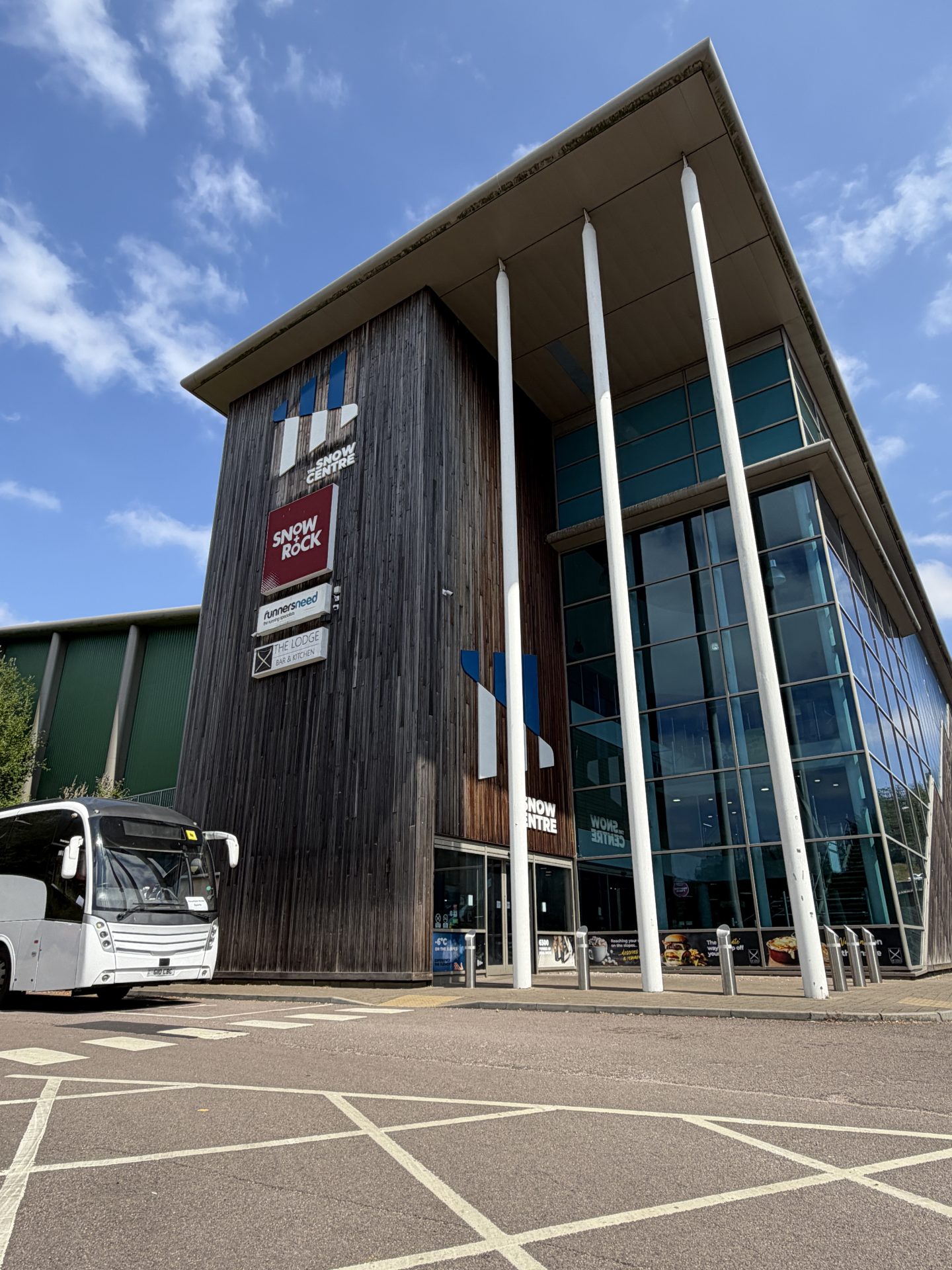 A modern building with tall white columns, a glass facade, and wooden panelling stands prominently in Hemel Hempstead. Signs for Snow+Rock and other businesses suggest its connection to The Snow Centre, the renowned indoor ski destination. A white coach is parked outside under a partly cloudy sky.