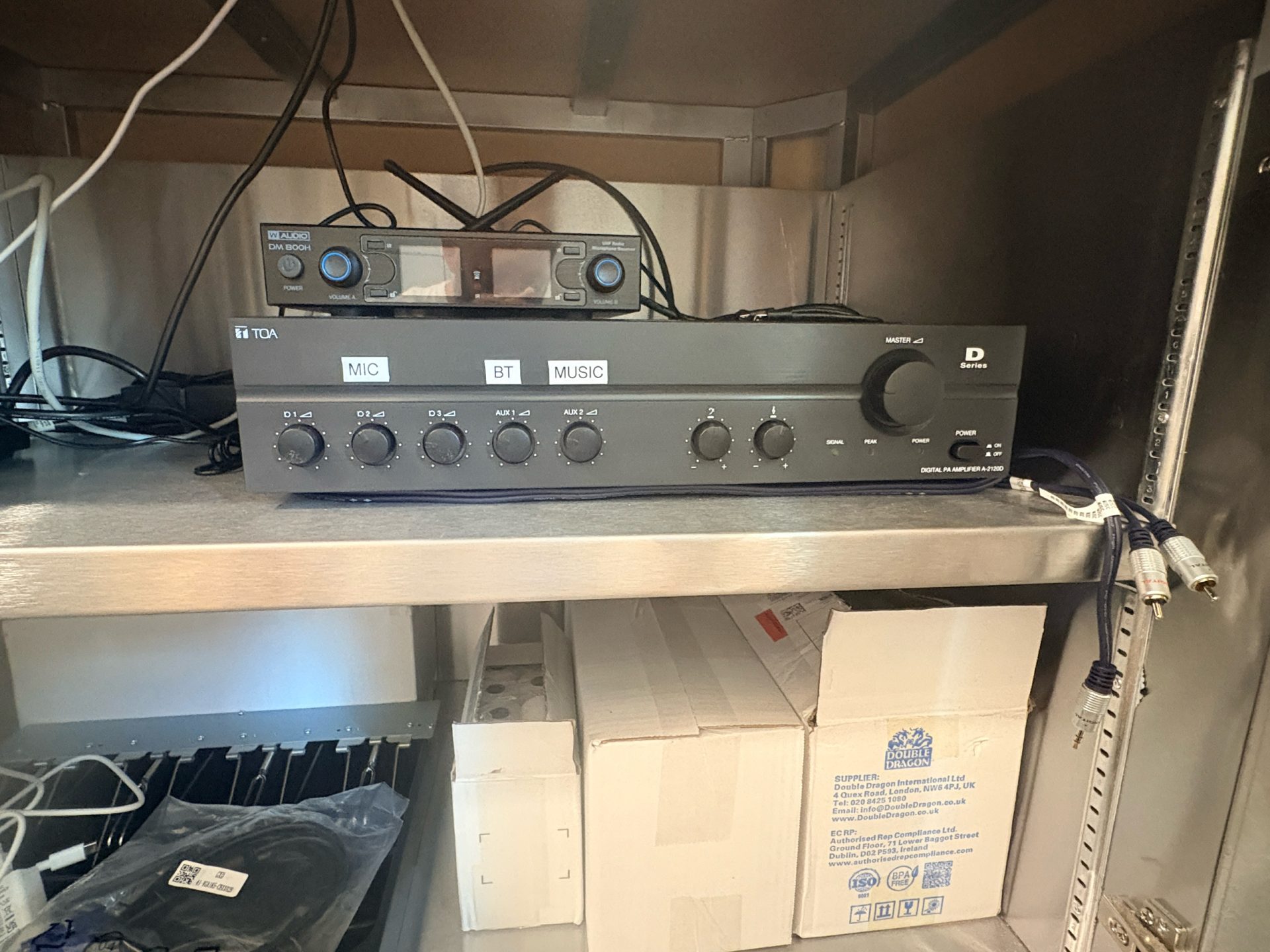 A black audio amplifier with labelled knobs sits on a metal shelf, surrounded by wires and boxes—perhaps tucked away in the back room of a Thurrock bistro or even at a community event for St Luke’s Hospice. The shelf below holds cables and more boxes.