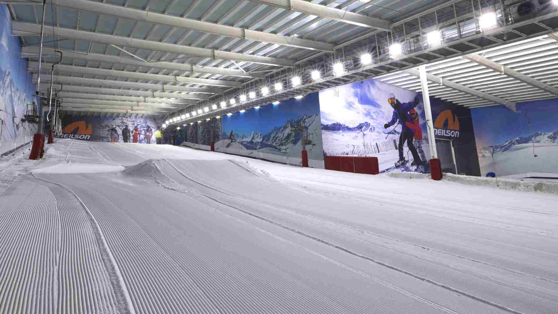 Indoor ski slope with artificial snow, groomed pistes, and colourful murals of snowy mountains and skiers on the walls. Bright ceiling lights illuminate the spacious, enclosed facility at the Snow Centre in Hemel Hempstead.