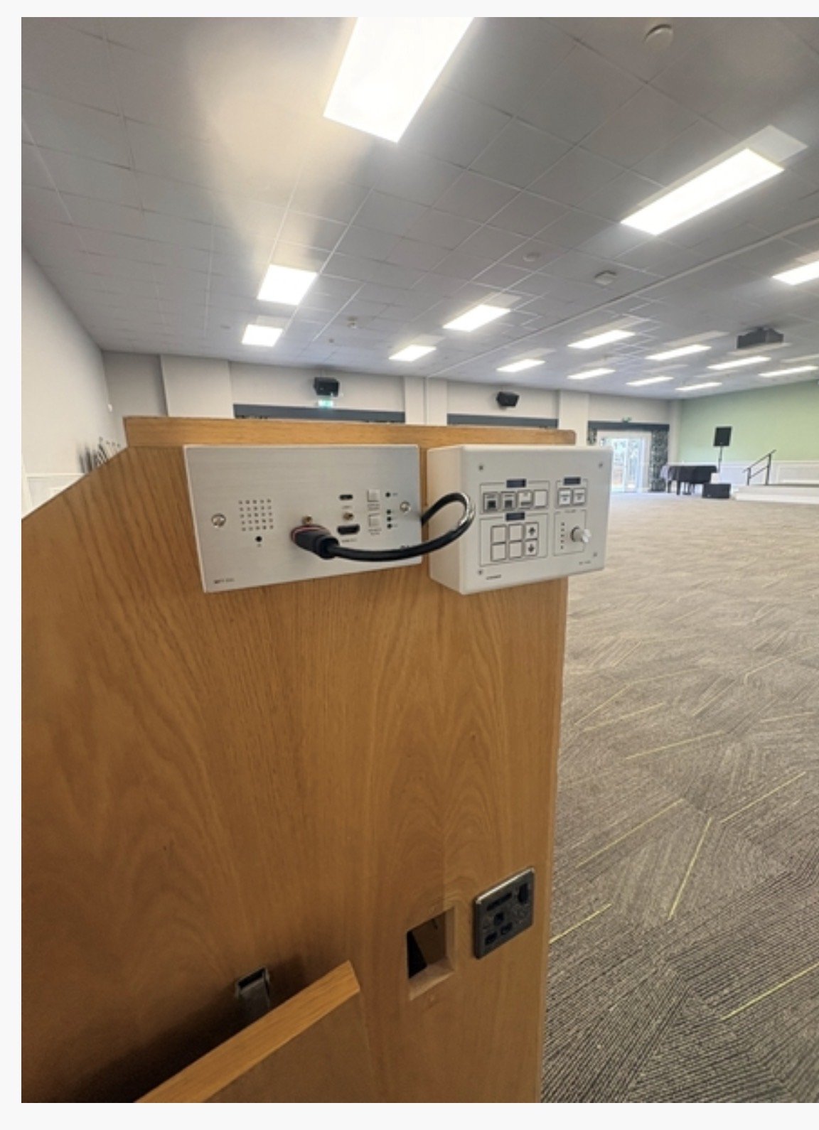 A wooden lectern with control panels and cables stands in an empty, well-lit conference room at Highgate House in Northampton, featuring carpeted floors and a green feature wall in the background.