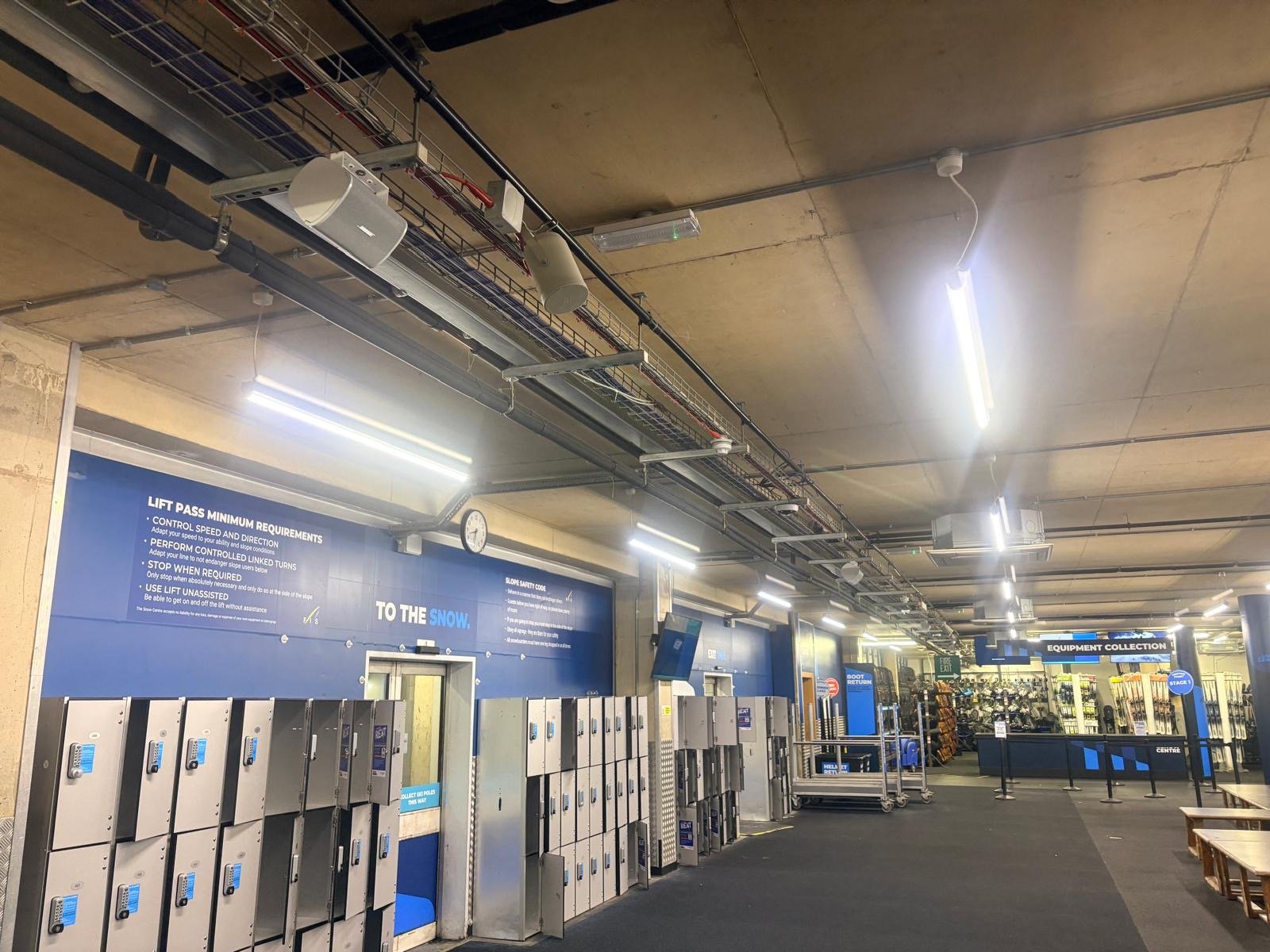 A brightly lit changing room with blue lockers, benches, and signs on the walls welcomes visitors to The Snow Centre in Hemel Hempstead. The equipment collection area is visible in the background, perfect for kitting up before enjoying some indoor skiing. Exposed pipes and lights hang from the ceiling, adding to the modern atmosphere.