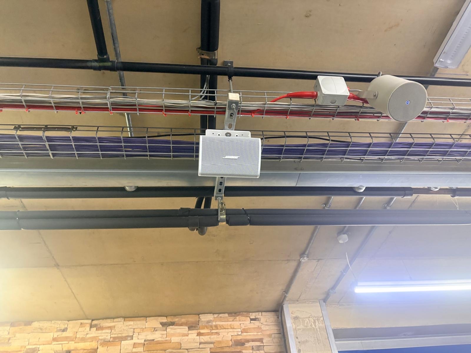 Ceiling-mounted speaker and round device attached to a metal cable tray, with various cables and pipes running along the ceiling at the Snow Centre in Hemel Hempstead. A brick-patterned wall and a fluorescent light are visible below.
