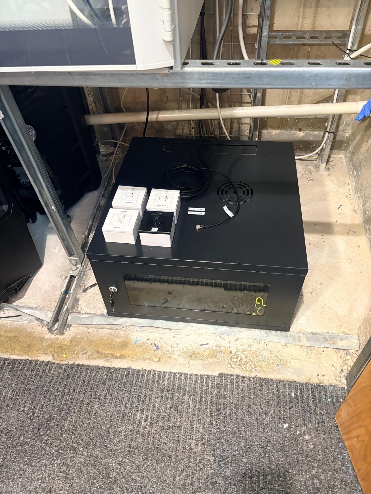 Black server cabinet on a concrete floor under a metal shelf, with three white boxes, a small black device, and coiled cables placed on top. Exposed pipes and wiring in the background evoke an industrial feel reminiscent of facilities near Hemel Hempstead’s Snow Centre.