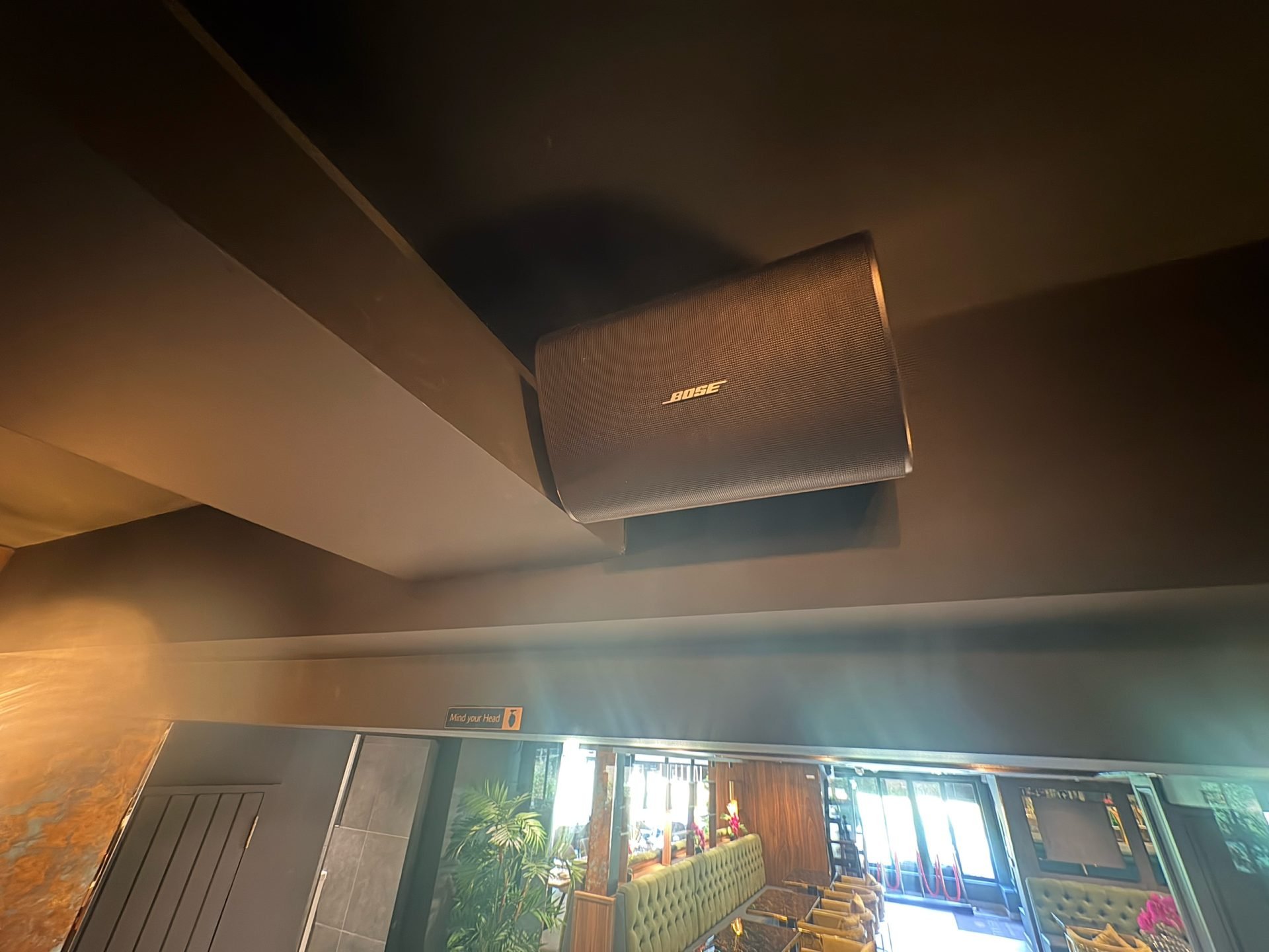 A black Bose speaker is mounted high on a dark ceiling inside Tanishq Restaurant, a modern room with seating, plants, and natural light from large windows in the background. A black Bose speaker is mounted high on a dark ceiling inside Tanishq Restaurant, a modern room with seating, plants, and natural light from large windows in the background.