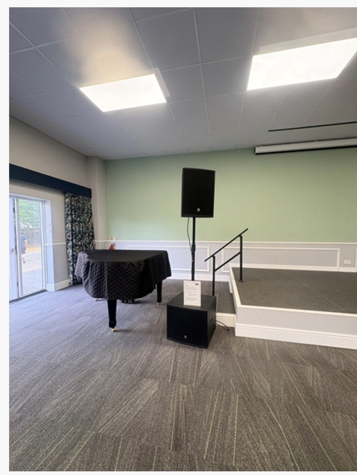 A small stage with a speaker, sign, and black-draped piano sits in a carpeted room with pale green walls, white trim, and large ceiling lights at Highgate House in Northampton. Sliding glass doors are on the left side.