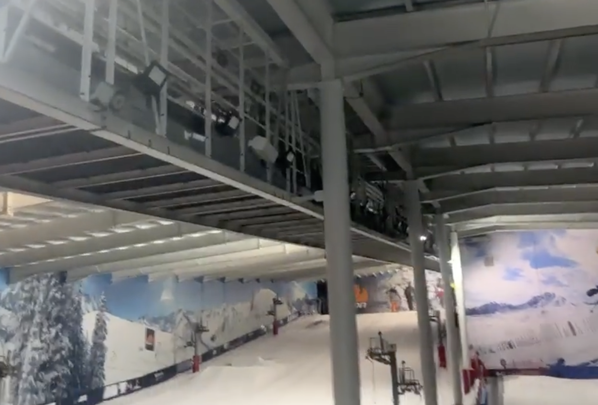 Indoor ski slope with artificial snow, steel beams overhead, and walls decorated with snowy mountain murals at The Snow Centre in Hemel Hempstead. Red and white ski barriers line the slope under bright lighting.