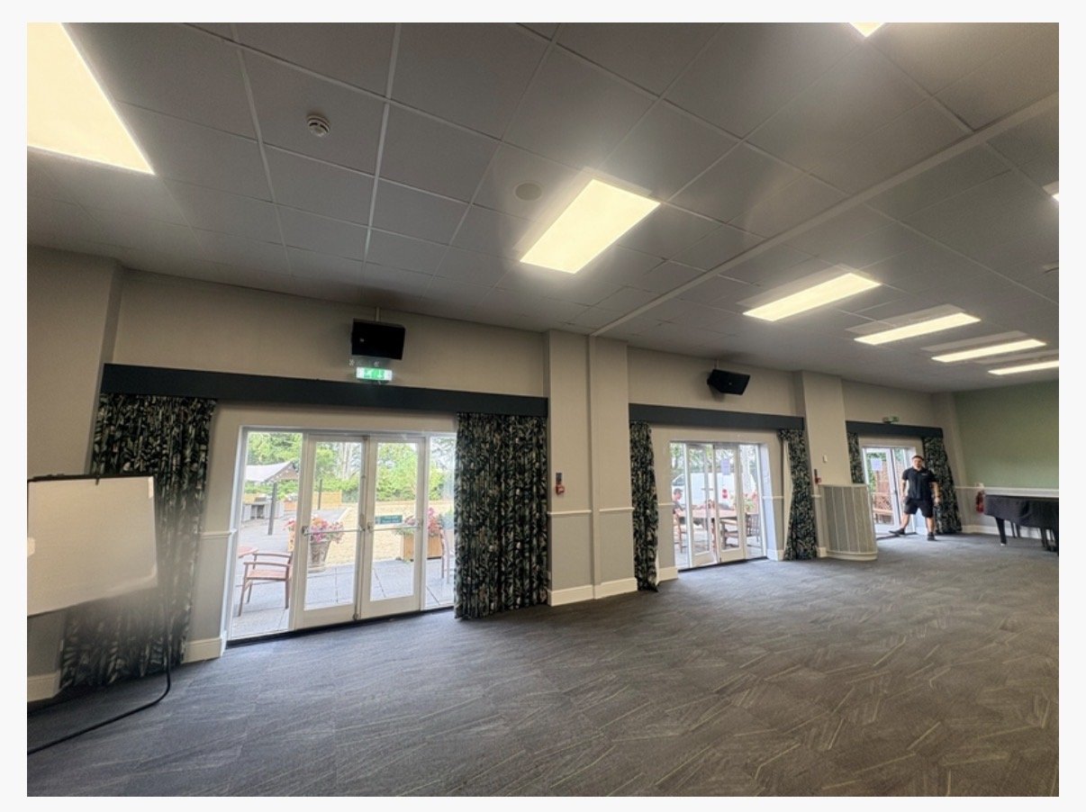 Spacious room at Highgate House in Northampton, with large windows and glass doors opening to a patio. The room has grey carpet, ceiling lights, floral curtains, a whiteboard, and a few people near the exit.