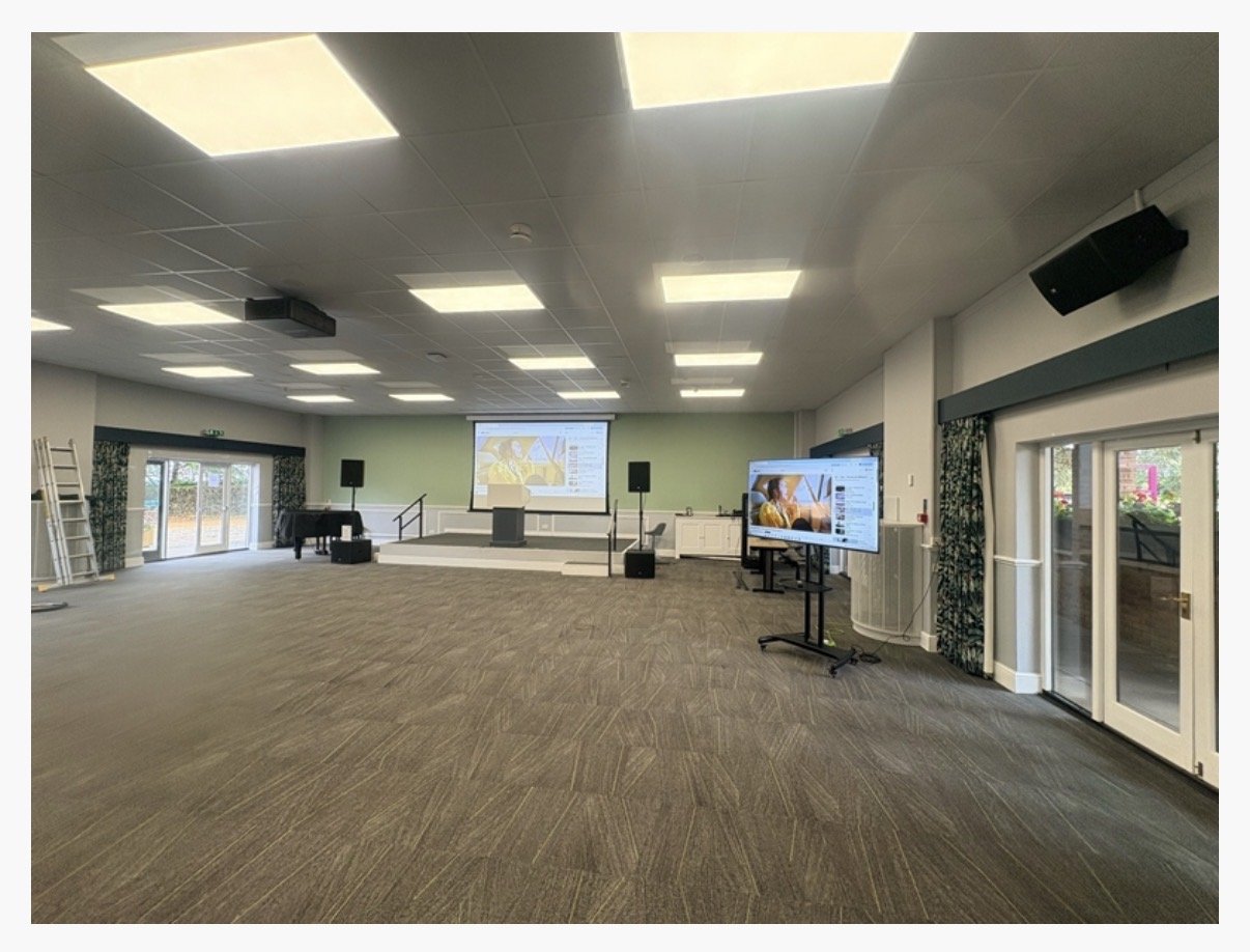 Spacious conference room at Highgate House in Northampton with brown carpet, large ceiling lights, a stage with screens and speakers, a TV display on a stand, and windows with curtains along the right wall.