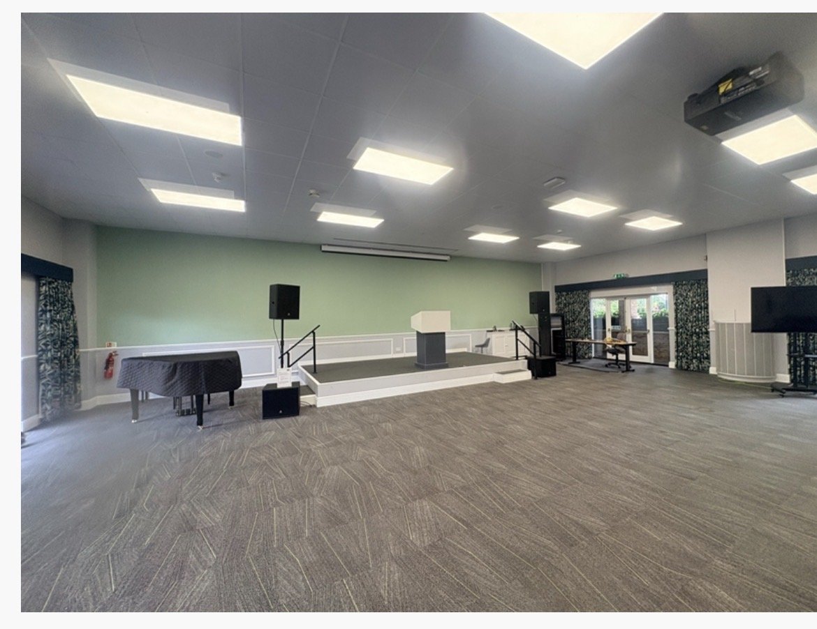 A spacious, well-lit room at Highgate House, Northampton, featuring a stage, lectern, piano, and audio equipment. The floor is carpeted, and large windows with curtains allow natural light in. There are ceiling lights and exit doors visible.