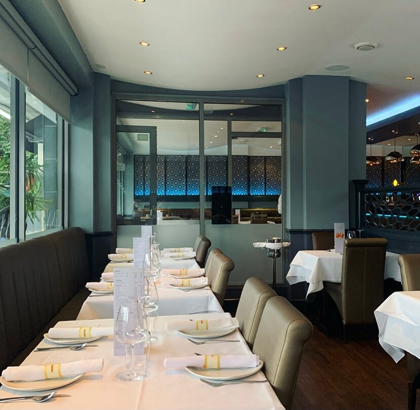 A modern restaurant dining area, Cinnamon Restaurant in North Weald, features neatly set tables with white tablecloths, rolled serviettes, wine glasses, and beige chairs, complemented by blue accent lighting and large windows that provide natural light. A modern restaurant dining area, Cinnamon Restaurant in North Weald, features neatly set tables with white tablecloths, rolled serviettes, wine glasses, and beige chairs, complemented by blue accent lighting and large windows that provide natural light.