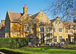 Large historic mansion with yellow stone walls, multiple chimneys, and gabled roofs, set in a well-kept garden with trees and outdoor seating on a sunny day—this elegant property evokes the charming character of Highgate House near Northampton.
