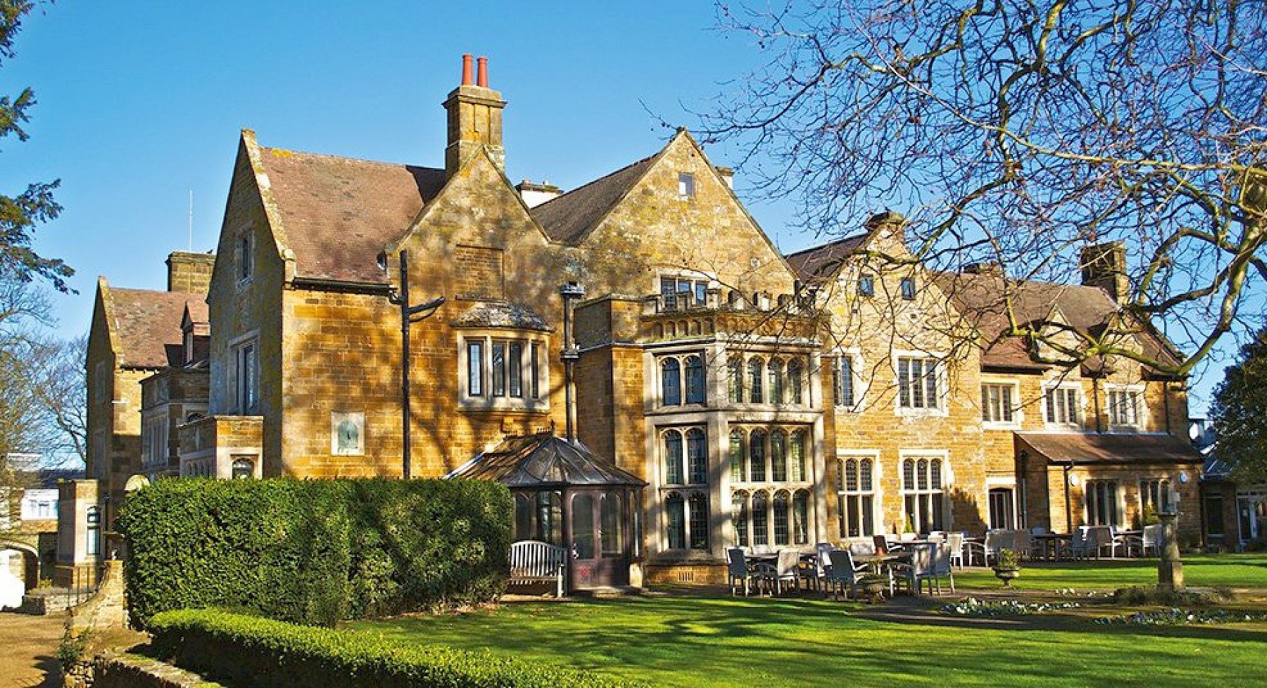 Large historic mansion with yellow stone walls, multiple chimneys, and gabled roofs, set in a well-kept garden with trees and outdoor seating on a sunny day—this elegant property evokes the charming character of Highgate House near Northampton.