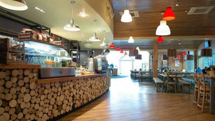 A modern café with wood accents, pendant lights, wooden tables, and chairs nestled in Hemel Hempstead. The counter features stacked wood logs, complemented by coffee machines and a display case behind it. The space appears bright and welcoming—an ideal spot to relax after a session at The Snow Centre or trying indoor skiing nearby.