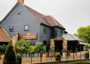 A rustic building houses Tanishq Restaurant & Bar in Toot Hill. The exterior is grey wood, with potted plants and flower arrangements by the entrance. Trees and a cloudy sky are in the background, creating a welcoming ambience for this charming restaurant.