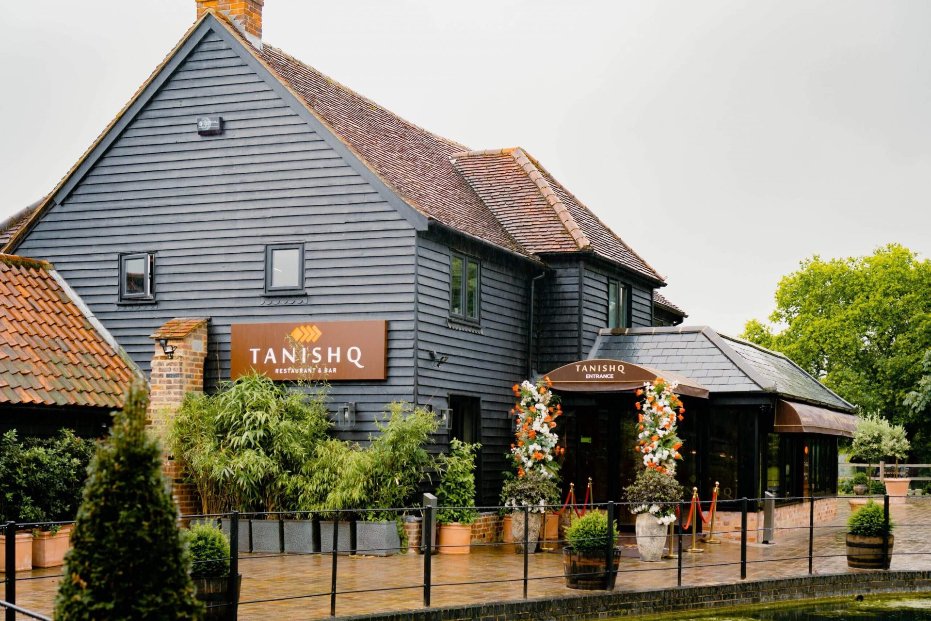 A rustic building houses Tanishq Restaurant & Bar in Toot Hill. The exterior is grey wood, with potted plants and flower arrangements by the entrance. Trees and a cloudy sky are in the background, creating a welcoming ambience for this charming restaurant. A rustic building houses Tanishq Restaurant & Bar in Toot Hill. The exterior is grey wood, with potted plants and flower arrangements by the entrance. Trees and a cloudy sky are in the background, creating a welcoming ambience for this charming restaurant.