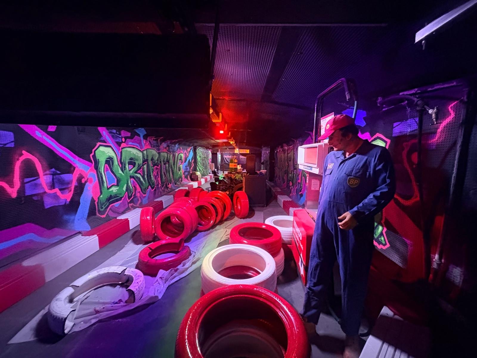 A person in a blue boiler suit stands near stacked red and white tyres in a dimly lit room with colourful graffiti on the walls and red overhead lighting, creating an atmosphere reminiscent of an underground London pub near London Bridge.