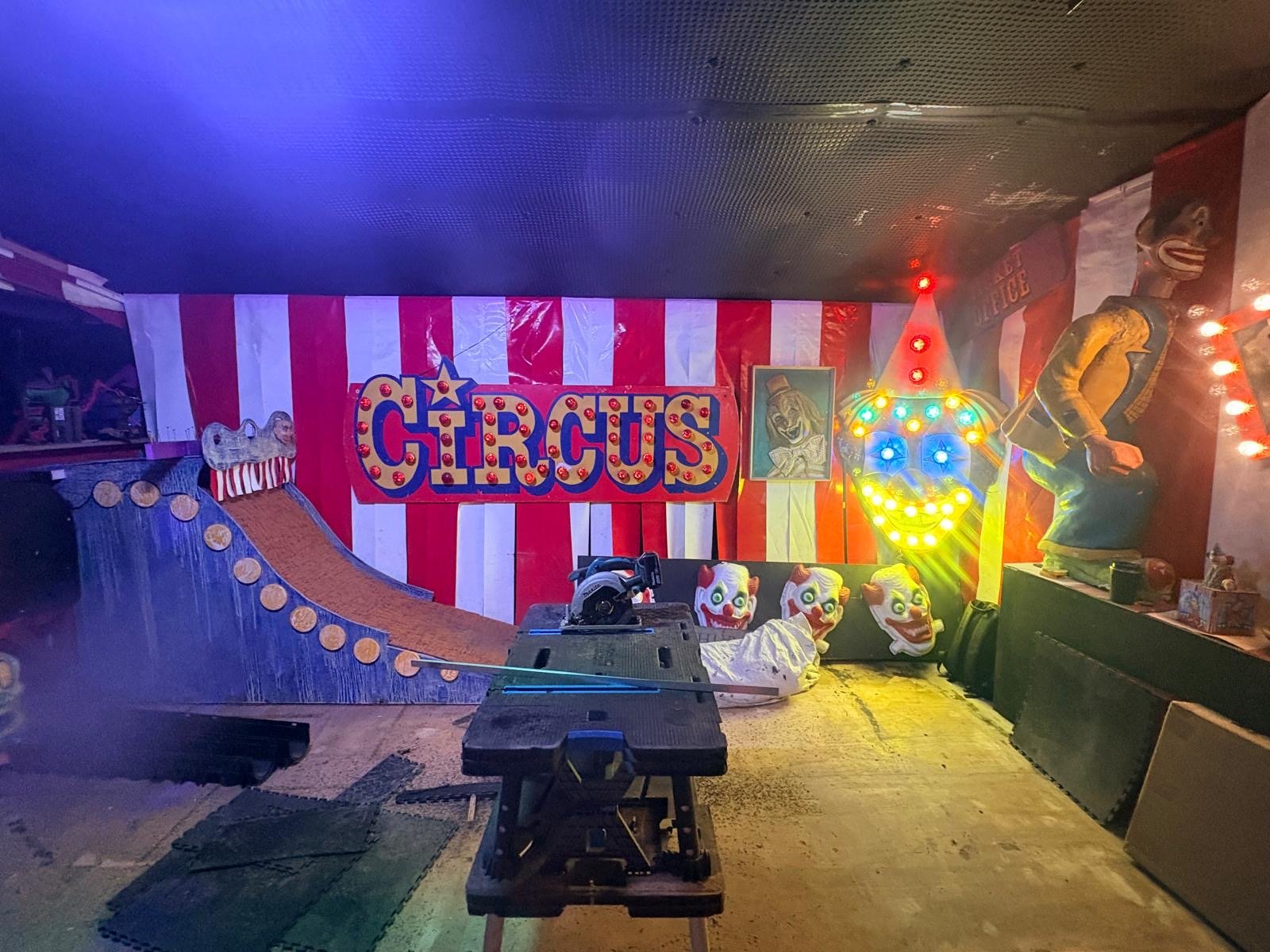 Room decorated as a circus with striped red and white walls, a large CIRCUS sign, clown props, a mini ramp, and bright clown face lights. Tools and pads are on the floor. The atmosphere is colourful and quirky—an experience reminiscent of the vibrant energy found in a Bierkeller near London Bridge.
