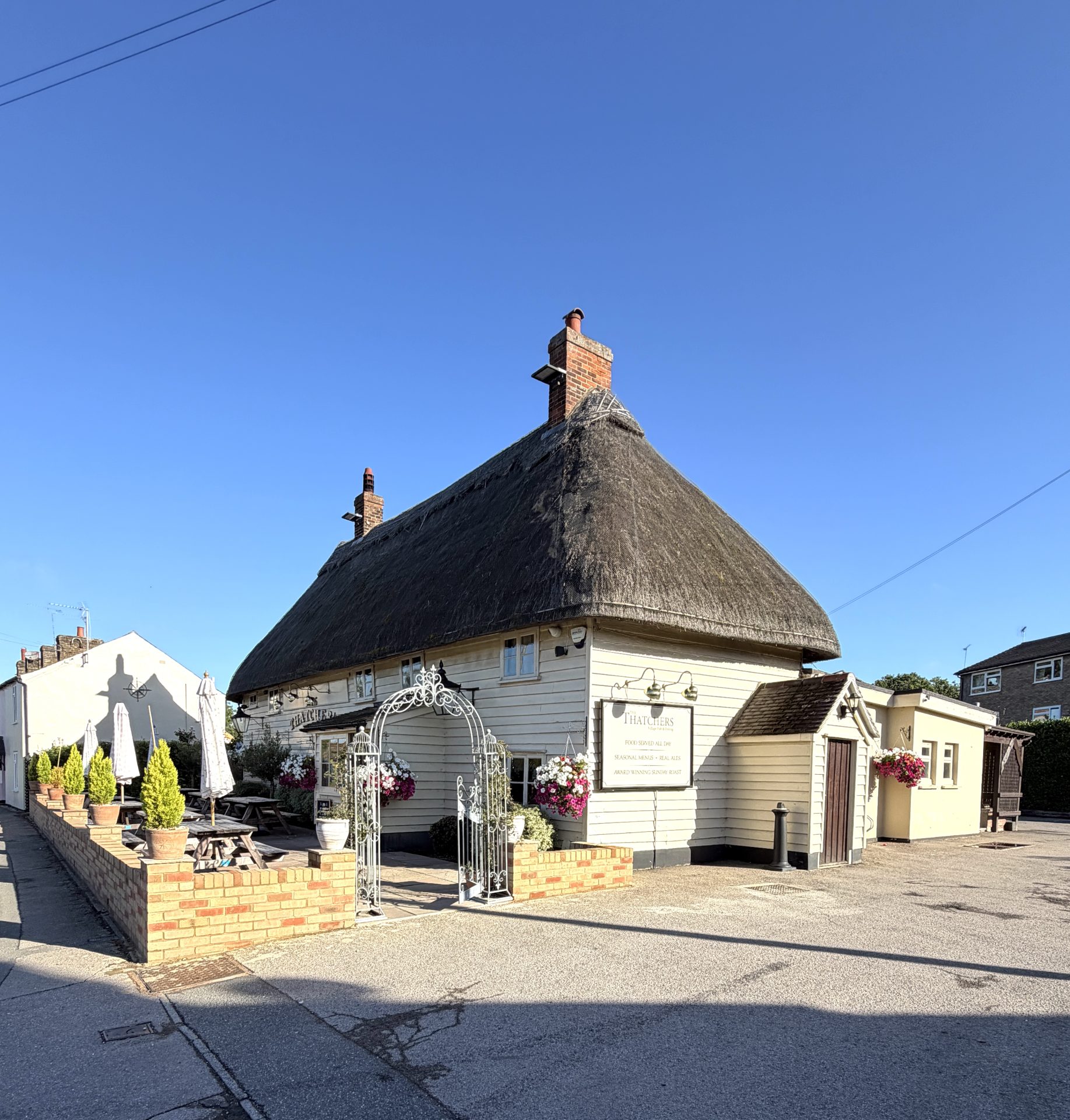A charming thatched-roof pub with white walls and brick accents sits under a clear blue sky in Hatfield Heath, featuring outdoor seating, hanging flower baskets, and a decorative metal arch at the entrance—a welcoming scene for visitors and Thatchers enthusiasts alike.