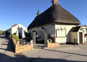 A thatched-roof pub, The Thatchers, with hanging flower baskets and an iron archway entrance sits on a sunny street in Hatfield Heath. There are outdoor tables with parasols on a raised patio beside the building.