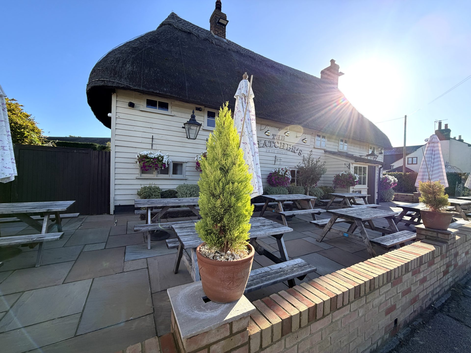 A thatched-roof pub, The Thatchers, sits at the heart of Hatfield Heath with outdoor wooden benches, potted plants, and parasols. The sun is shining brightly, casting long shadows on the patio.