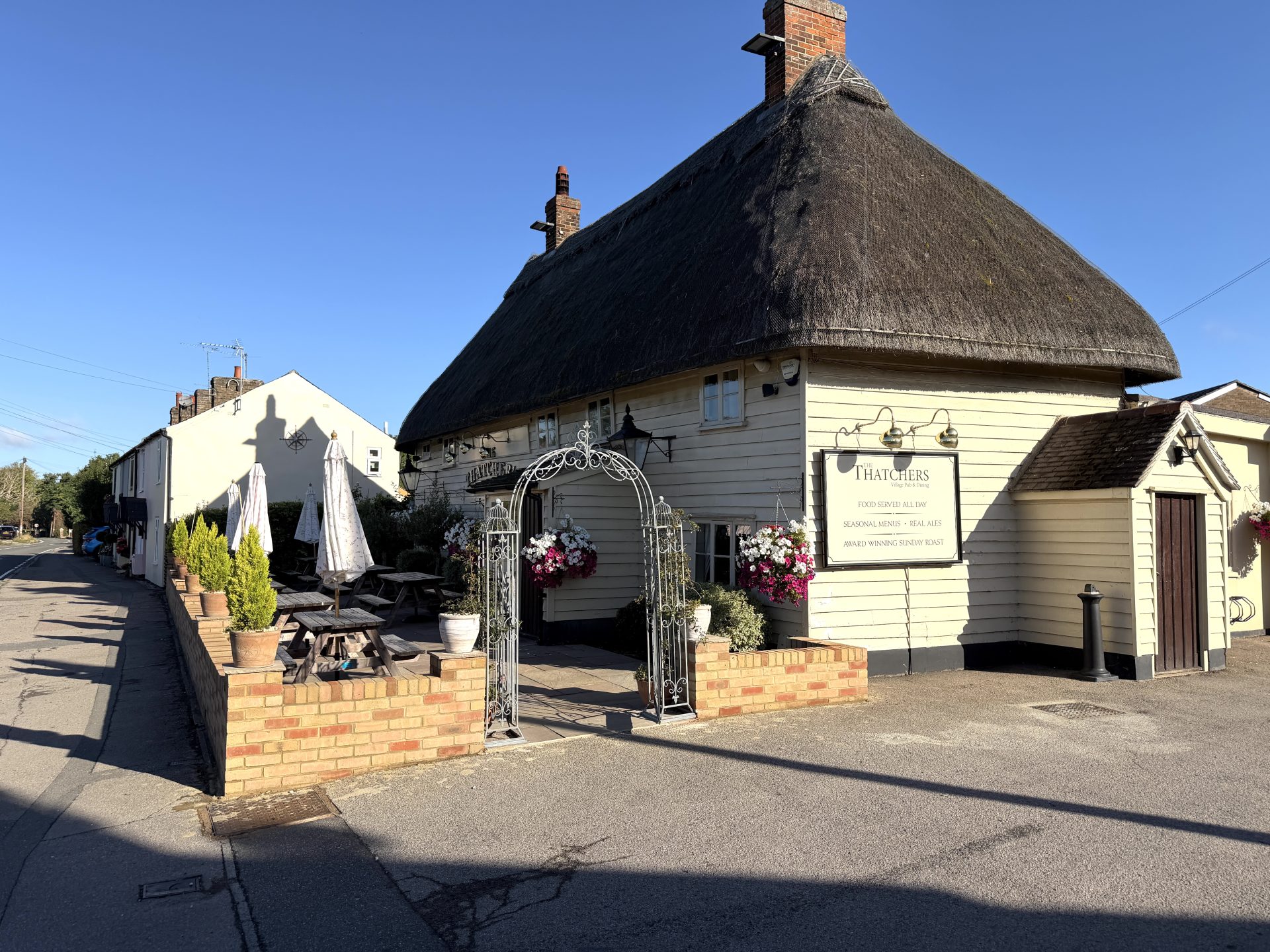 A thatched-roof pub, The Thatchers, with hanging flower baskets and an iron archway entrance sits on a sunny street in Hatfield Heath. There are outdoor tables with parasols on a raised patio beside the building.