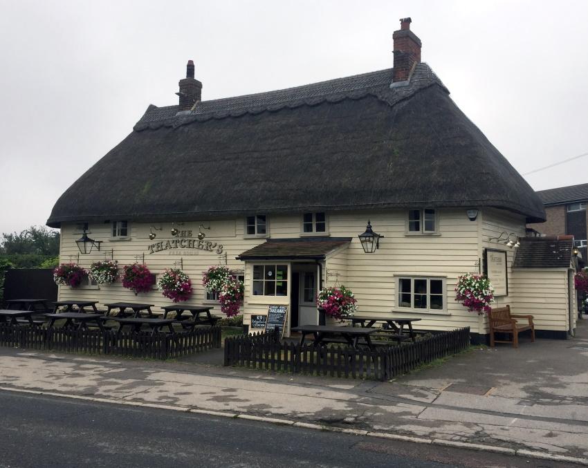 A thatched-roof pub called The Thatchers, with cream cladding, black trim, hanging flower baskets, wooden benches, and a small sign out front, sits on Hatfield Heath, seen on a cloudy day from across a quiet road.