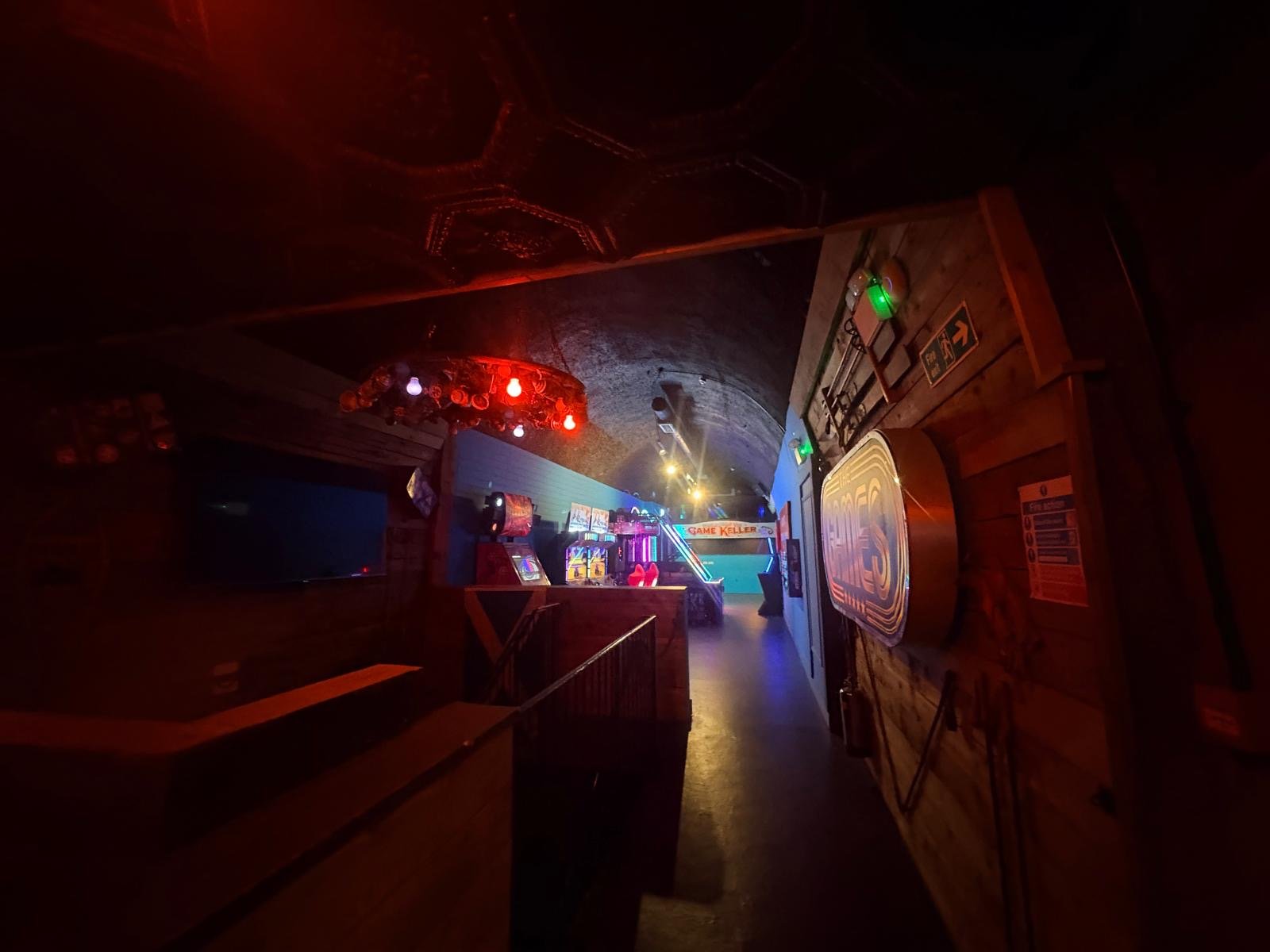 A dimly lit arcade with colourful lights, vintage arcade machines, a pool table in the distance, and wooden walls adorned with signs and decorations—a hidden gem reminiscent of a classic London pub near London Bridge.